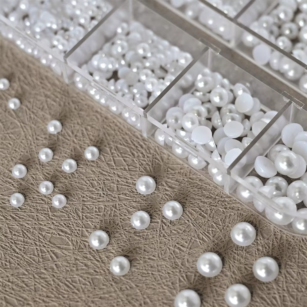 A transparent plastic container with multiple compartments, filled with small, round, glossy pearl beads in various sizes, with some beads scattered on a textured surface.