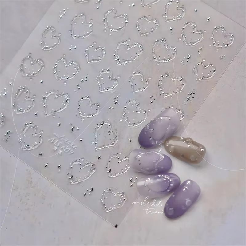 A sheet of nail stickers with heart shapes, small stars, and moons, alongside artificial nails painted in a gradient of purple and beige colors.