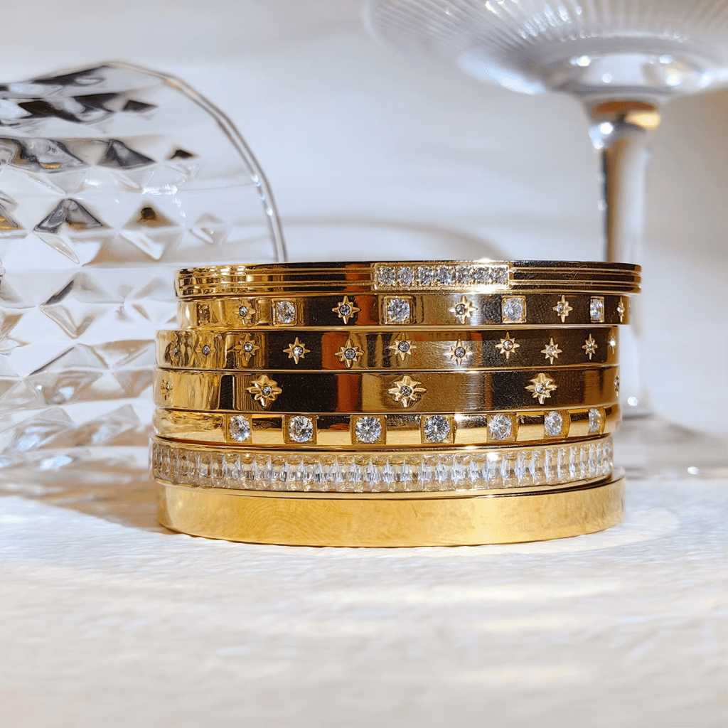 A stack of gold bracelets adorned with various designs and gemstones next to a crystal glass and a wine glass.