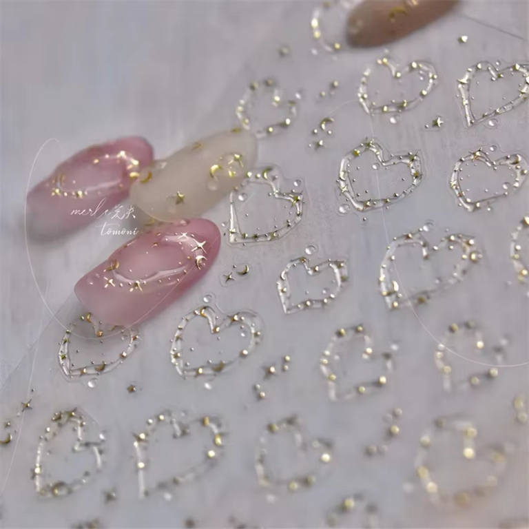 Pink and white gradient nails with small golden stars and crescent moon designs, alongside a sheet of transparent stickers with heart shapes and golden dots.