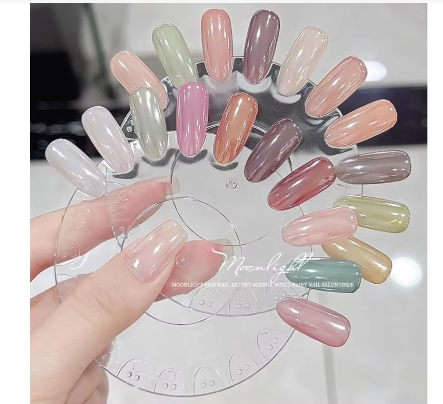 A clear plastic nail polish display wheel held by a hand, featuring 18 different nail polish colors ranging from light pastels to darker shades, including pinks, greens, browns, and purples.
