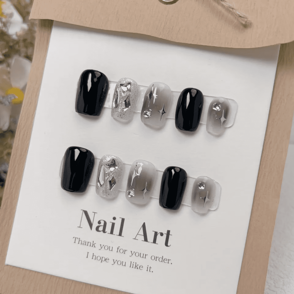 A set of artificial nails on a card labeled "Nail Art," featuring a mix of designs including solid black nails, silver glitter nails, and clear nails with silver embellishments and star patterns.
