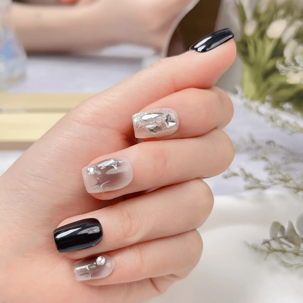 A close-up of a hand with a manicure featuring different nail designs, including solid black nails, glittery silver nails with metallic star and moon embellishments, and clear nails with silver star designs and small metallic studs.